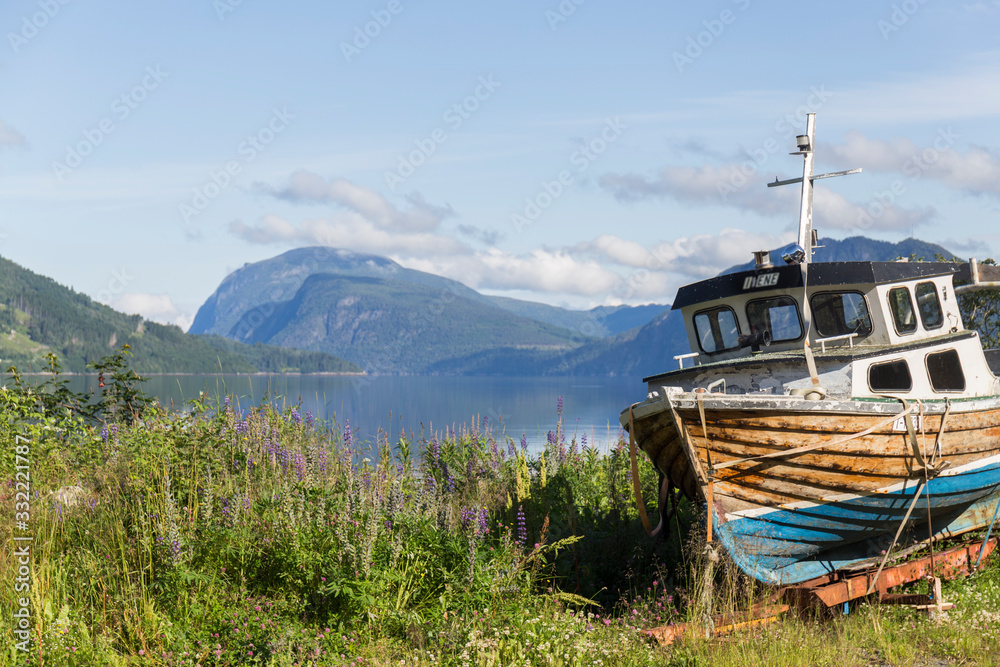 old wooden boat, brown, blue and white washed in the grass, next to a deep blue Norwegian fjord, in the background we can see the mountains, clear summer day with some clouds old wooden boat, brown, blue and white washed in the grass, next to a deep blue Norwegian fjord, in the background we can see the mountains, clear summer day with some clouds