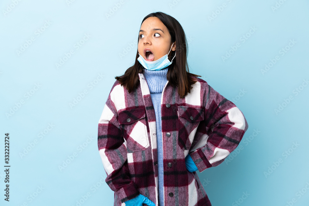 Young brunette mixed race woman protecting from the coronavirus with a mask and gloves over isolated blue background doing surprise gesture while looking to the side Young brunette mixed race woman protecting from the coronavirus with a mask and gloves over isolated blue background doing surprise gesture while looking to the side