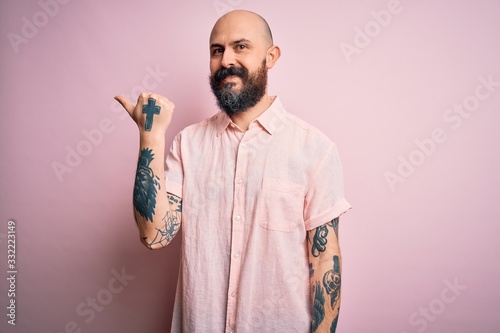 Handsome bald man with beard and tattoo wearing casual shirt over isolated pink background smiling with happy face looking and pointing to the side with thumb up.