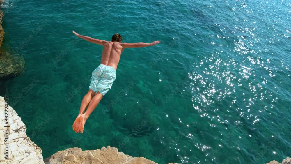 SLOW MOTION, COPY SPACE: Young man on a relaxing summer vacation in Croatia does cliff diving on a sunny day. Cinematic shot of a man jumping off a rocky ledge and into the glistening blue ocean.
