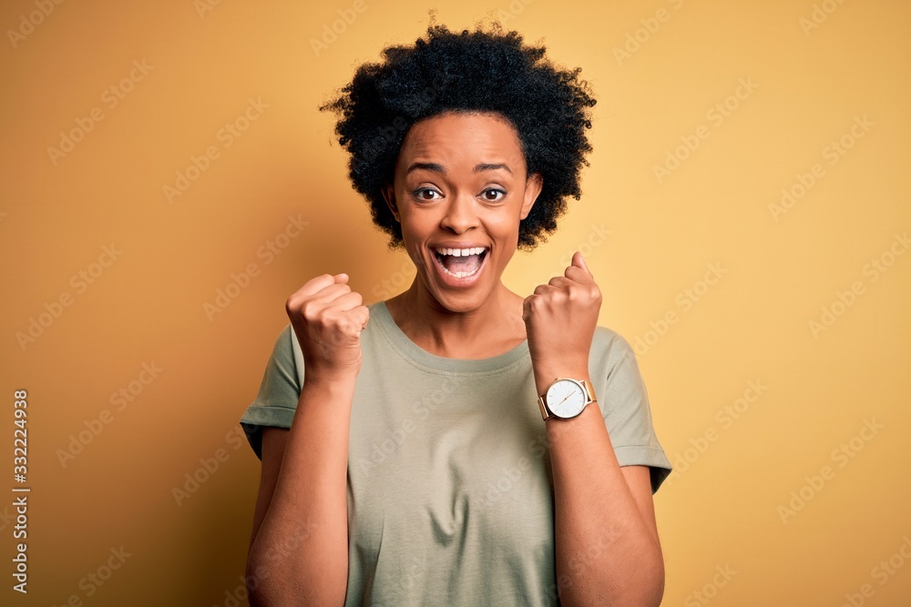 Young beautiful African American afro woman with curly hair wearing casual t-shirt celebrating surprised and amazed for success with arms raised and open eyes. Winner concept. Young beautiful African American afro woman with curly hair wearing casual t-shirt celebrating surprised and amazed for success with arms raised and open eyes. Winner concept.