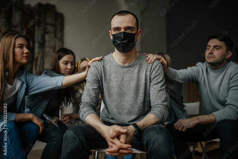 ill caucasian guy in medical protective mask suffer from incurable virus infection, sit in center of isolated room with strangers supporting him in such hard time. sickness, cancer, coronavirus