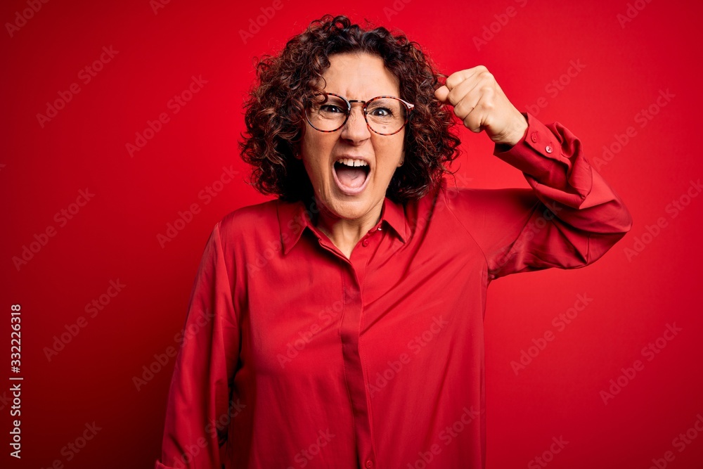 Middle age beautiful curly hair woman wearing casual shirt and glasses over red background angry and mad raising fist frustrated and furious while shouting with anger. Rage and aggressive concept.