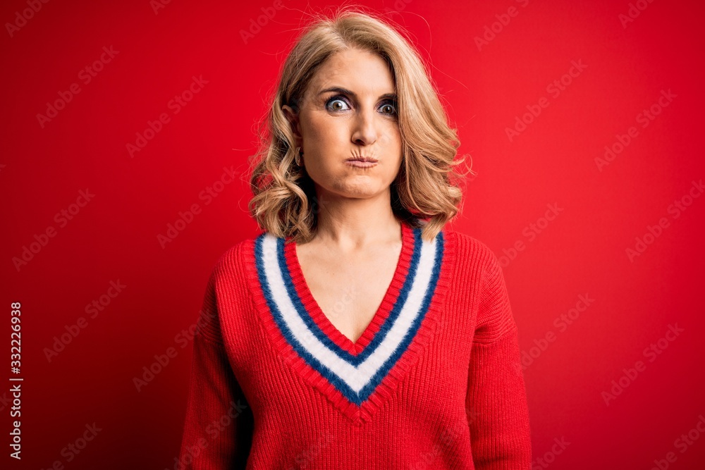 Middle age beautiful blonde woman wearing casual sweater over isolated red background puffing cheeks with funny face. Mouth inflated with air, crazy expression. Middle age beautiful blonde woman wearing casual sweater over isolated red background puffing cheeks with funny face. Mouth inflated with air, crazy expression.