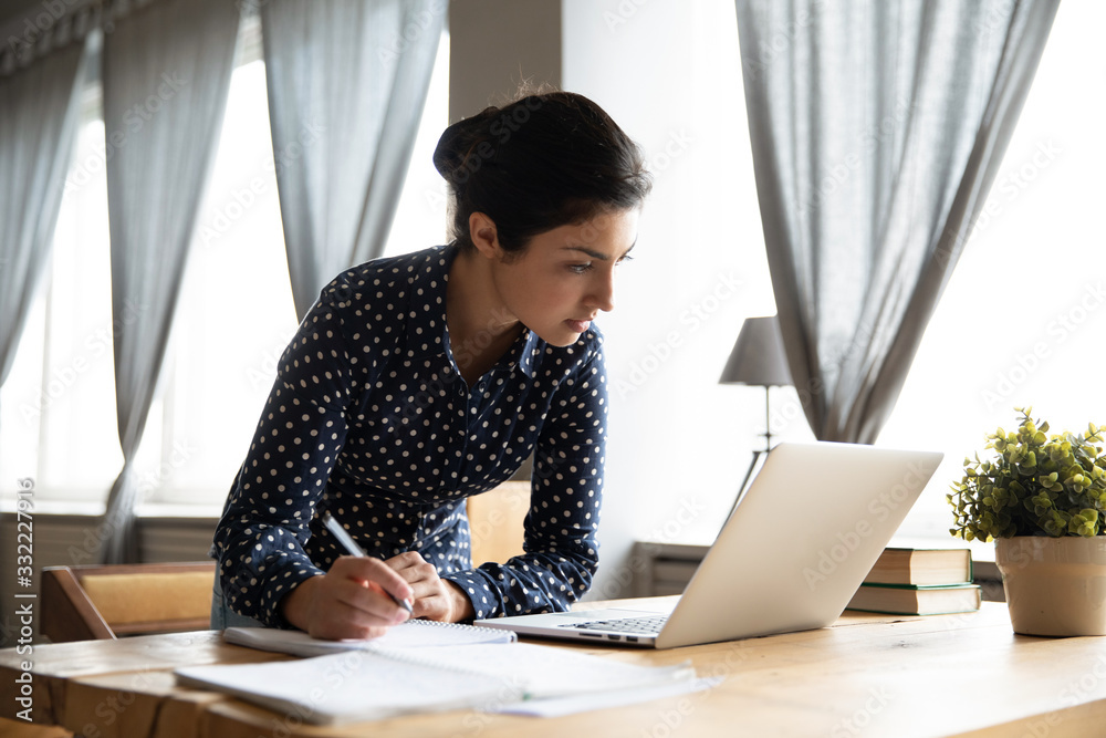 Busy Indian woman standing at table, writing notes in notebook with pen,  focused female student using laptop, studying online at home, looking at  screen, reading important information Stock Photo | Adobe Stock