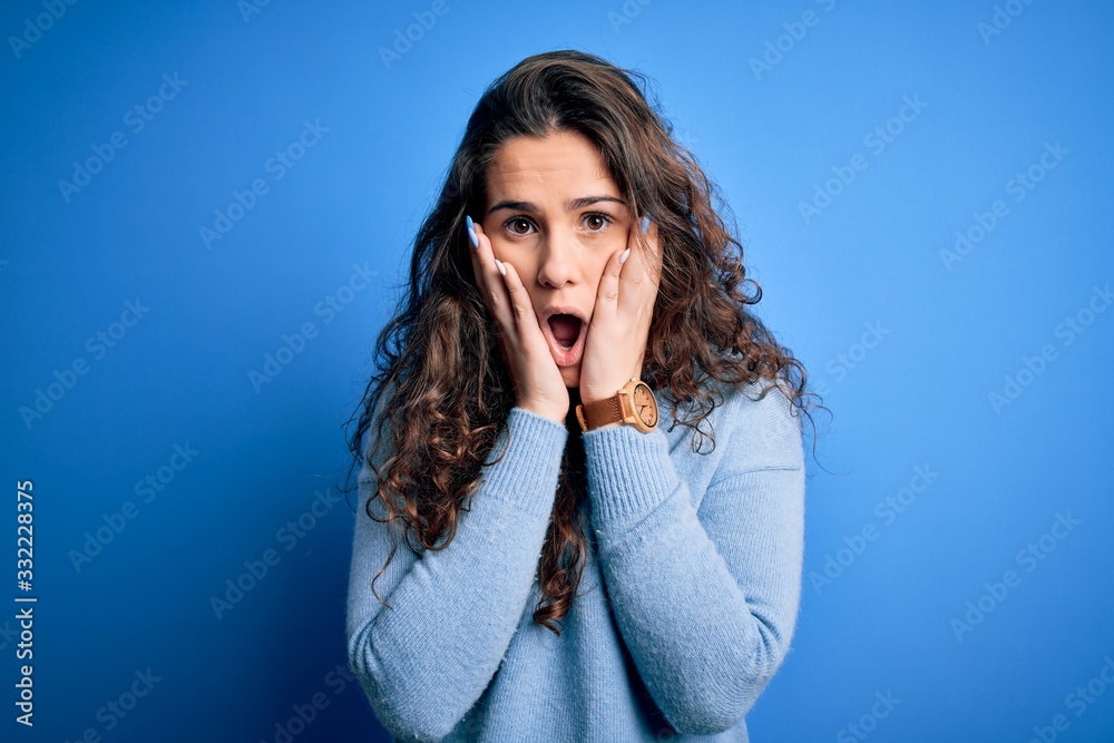 Young beautiful woman with curly hair wearing blue casual sweater over isolated background afraid and shocked, surprise and amazed expression with hands on face Young beautiful woman with curly hair wearing blue casual sweater over isolated background afraid and shocked, surprise and amazed expression with hands on face