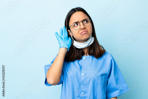 Surgeon woman over isolated blue background listening to something by putting hand on the ear