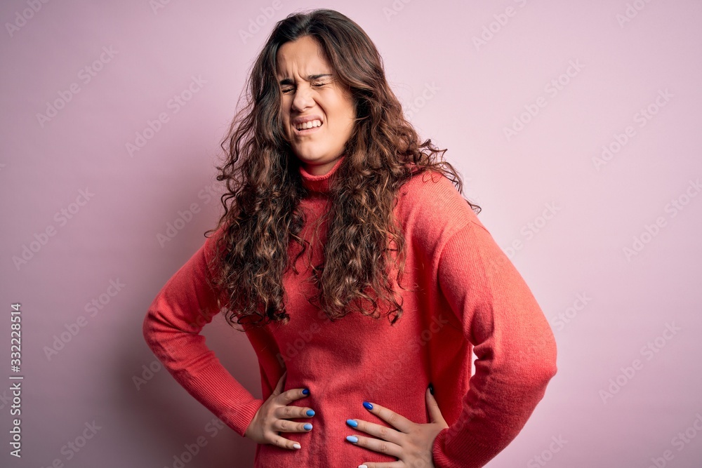 Young beautiful woman with curly hair wearing turtleneck sweater over pink background with hand on stomach because nausea, painful disease feeling unwell. Ache concept. Young beautiful woman with curly hair wearing turtleneck sweater over pink background with hand on stomach because nausea, painful disease feeling unwell. Ache concept.