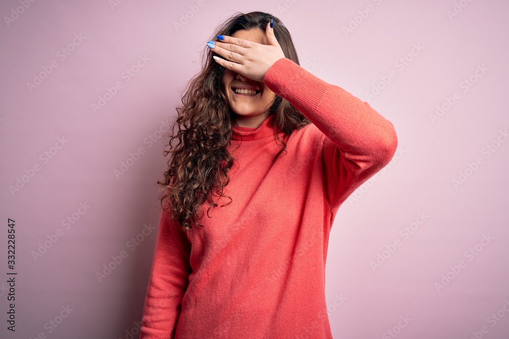 Young beautiful woman with curly hair wearing turtleneck sweater over pink background smiling and laughing with hand on face covering eyes for surprise. Blind concept. Young beautiful woman with curly hair wearing turtleneck sweater over pink background smiling and laughing with hand on face covering eyes for surprise. Blind concept.