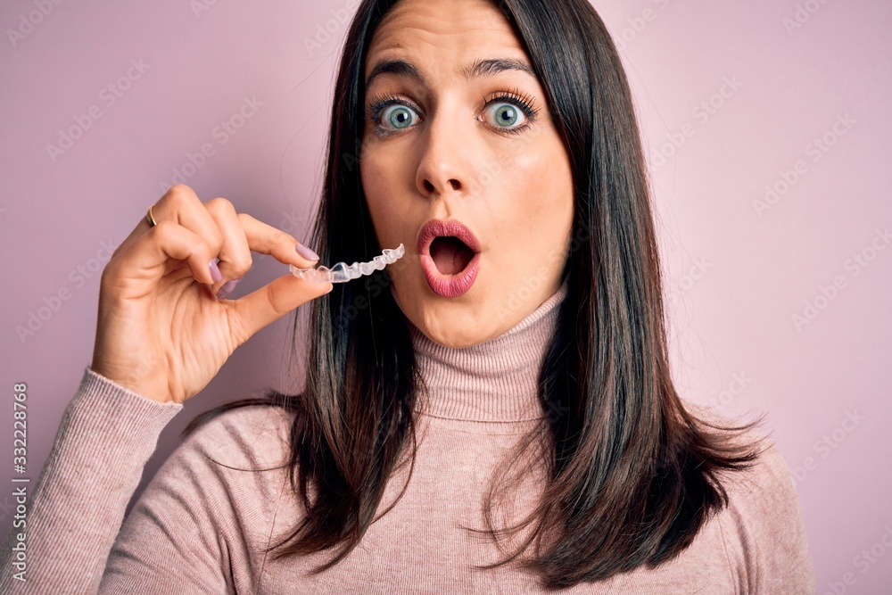 Young woman with blue eyes holding clear aligner standing over pink background scared in shock with a surprise face, afraid and excited with fear expression Young woman with blue eyes holding clear aligner standing over pink background scared in shock with a surprise face, afraid and excited with fear expression