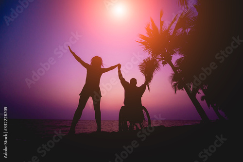 Disabled man in a wheelchair with his wife on the beach. Silhouettes at sunset