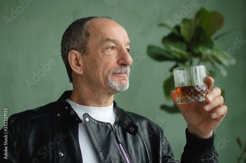 Male portrait of senior man in black leather jacket with glass of brandy indoors in loft style room with light green walls
