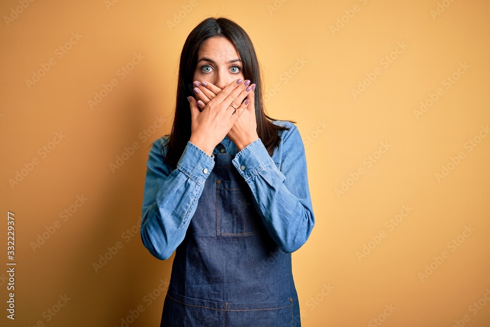 Young baker woman with blue eyes wearing apron standing over isolated yellow background shocked covering mouth with hands for mistake. Secret concept. Young baker woman with blue eyes wearing apron standing over isolated yellow background shocked covering mouth with hands for mistake. Secret concept.