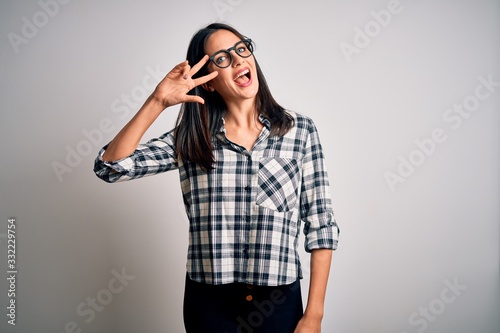 Young brunette woman with blue eyes wearing casual shirt and glasses over white background Doing peace symbol with fingers over face, smiling cheerful showing victory