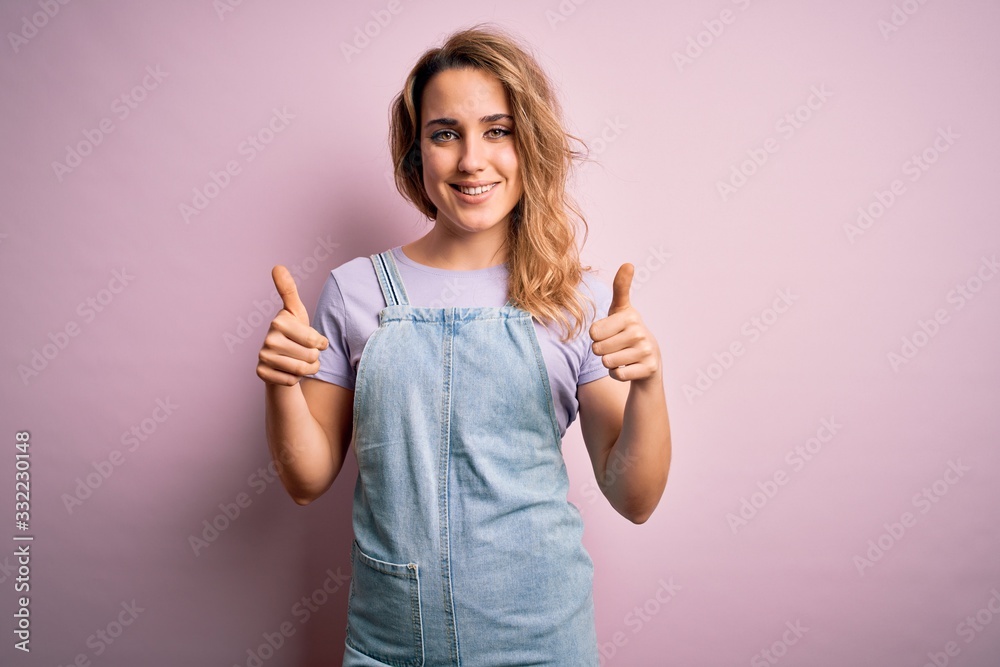 Young beautiful blonde woman wearing casual denim overalls standing over pink background success sign doing positive gesture with hand, thumbs up smiling and happy. Cheerful expression and winner