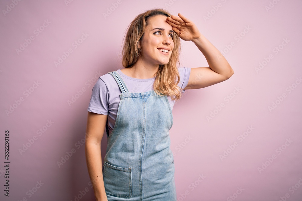 Young beautiful blonde woman wearing casual denim overalls standing over pink background very happy and smiling looking far away with hand over head. Searching concept.
