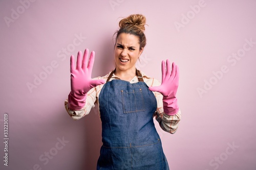 Young beautiful blonde cleaner woman doing housework wearing arpon and gloves afraid and terrified with fear expression stop gesture with hands, shouting in shock. Panic concept.