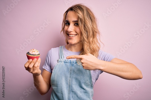 Young beautiful blonde woman eatimg chocolate cupcake over isolated pink background very happy pointing with hand and finger photo