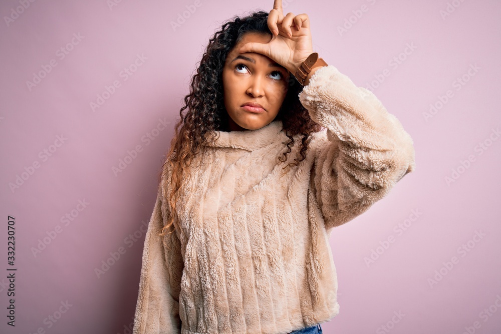 Young beautiful woman with curly hair wearing casual sweater standing over pink background making fun of people with fingers on forehead doing loser gesture mocking and insulting. Young beautiful woman with curly hair wearing casual sweater standing over pink background making fun of people with fingers on forehead doing loser gesture mocking and insulting.