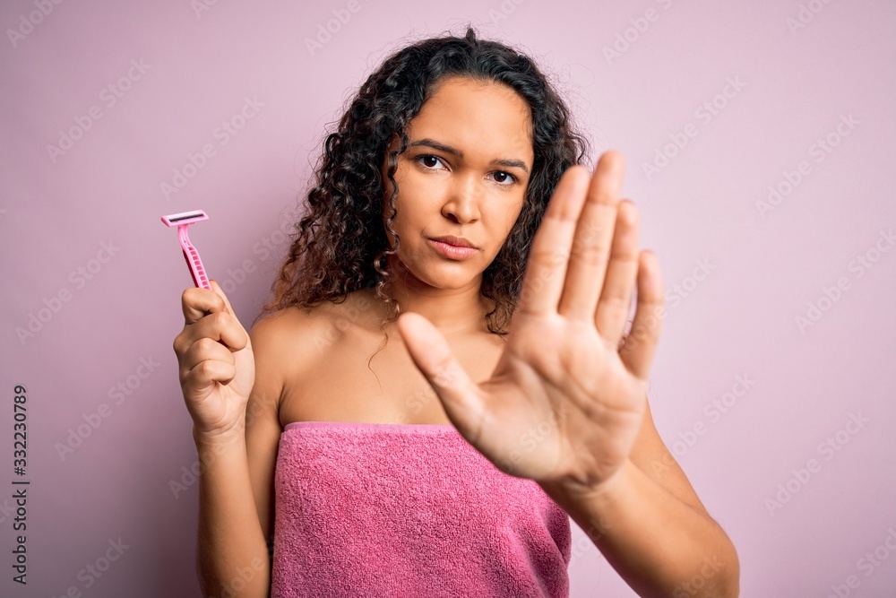 Young beautiful woman with curly hair wearing shower towel holding depilation razor with open hand doing stop sign with serious and confident expression, defense gesture Young beautiful woman with curly hair wearing shower towel holding depilation razor with open hand doing stop sign with serious and confident expression, defense gesture