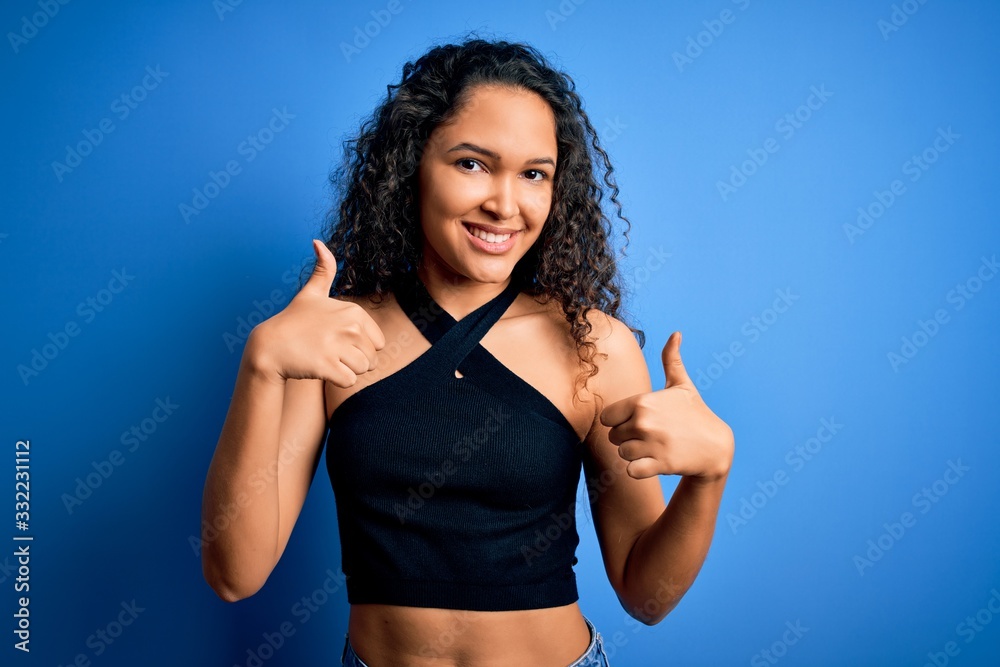 Young beautiful woman with curly hair wearing casual t-shirt standing over blue background success sign doing positive gesture with hand, thumbs up smiling and happy. Cheerful expression and winner Young beautiful woman with curly hair wearing casual t-shirt standing over blue background success sign doing positive gesture with hand, thumbs up smiling and happy. Cheerful expression and winner