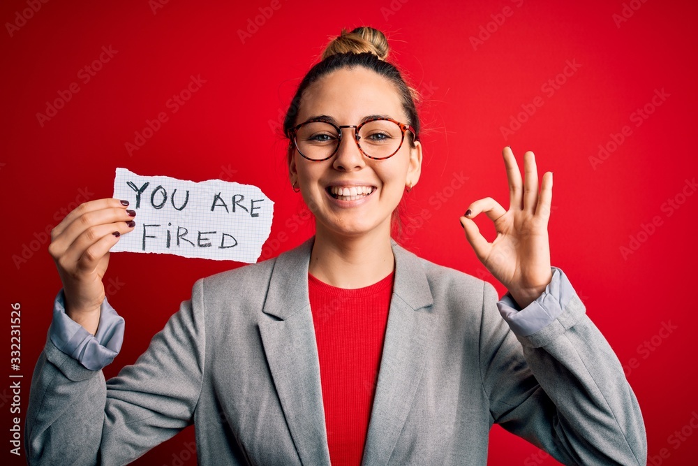 Young beautiful blonde woman with blue eyes holding paper with you are fired message doing ok sign with fingers, excellent symbol