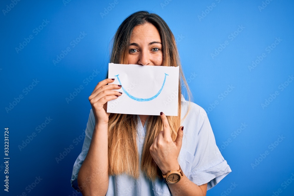 Young beautiful brunette woman holding card with smile illustration on mouth very happy pointing with hand and finger Young beautiful brunette woman holding card with smile illustration on mouth very happy pointing with hand and finger