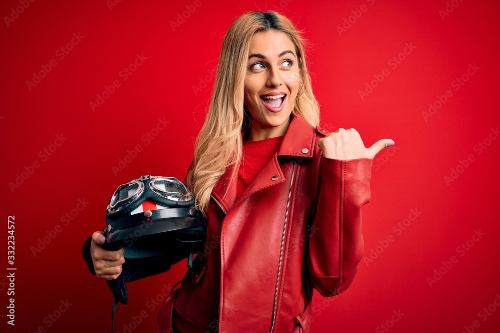 Naklejka premium Young beautiful blonde motorcyclist woman holding moto helmet over isolated red background pointing and showing with thumb up to the side with happy face smiling