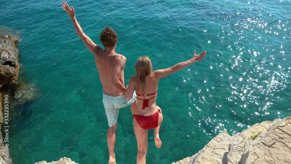 SLOW MOTION: Young couple holding hands jumps off a cliff and into the ocean. Cheerful male and female tourists hold hands while diving into the deep blue sea. Carefree man and woman jump into ocean.