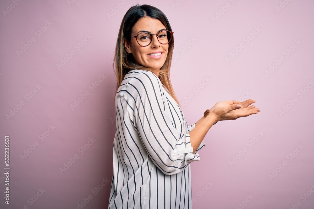 Young beautiful woman wearing casual striped t-shirt and glasses over pink background pointing aside with hands open palms showing copy space, presenting advertisement smiling excited happy Young beautiful woman wearing casual striped t-shirt and glasses over pink background pointing aside with hands open palms showing copy space, presenting advertisement smiling excited happy