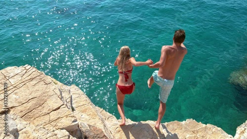 SLOW MOTION: Joyful tourist couple decides to jump off a rocky cliff and dive into sea. Unrecognizable man holds his girlfriend's hand while diving into the deep blue sea on a sunny summer day.