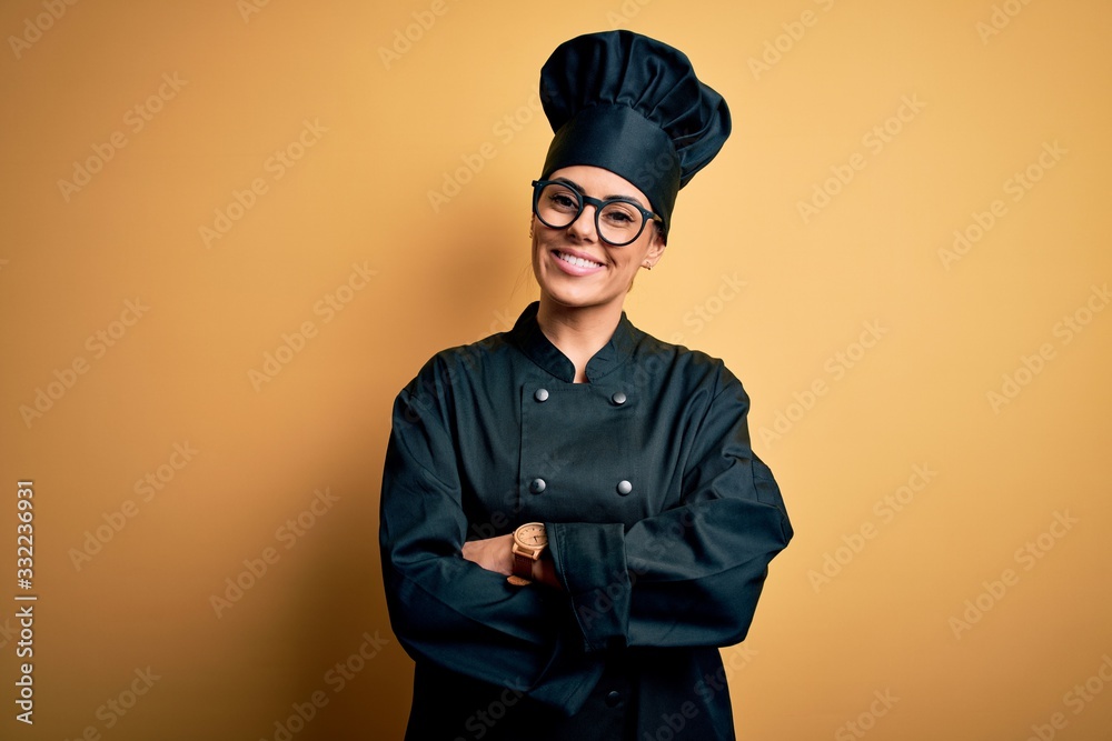 Young beautiful brunette chef woman wearing cooker uniform and hat over yellow background happy face smiling with crossed arms looking at the camera. Positive person.