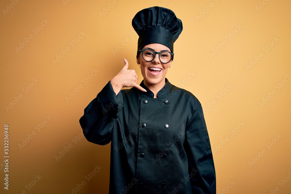 Young beautiful brunette chef woman wearing cooker uniform and hat over yellow background smiling doing phone gesture with hand and fingers like talking on the telephone. Communicating concepts. Young beautiful brunette chef woman wearing cooker uniform and hat over yellow background smiling doing phone gesture with hand and fingers like talking on the telephone. Communicating concepts.