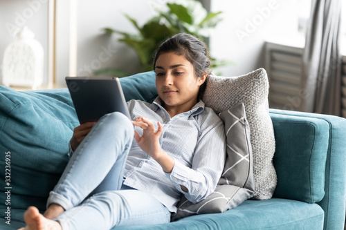 Peaceful Indian woman using computer tablet, relaxing on couch, chatting in social network or shopping online, playing game, focused young female looking at screen, reading book, lying on cozy sofa