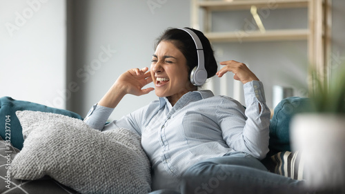Excited Indian woman wearing wireless headphones singing popular song, having fun, happy smiling young female listening to favorite music, spending weekend, relaxing on cozy couch in living room
