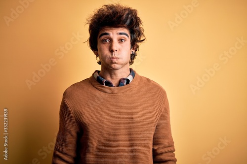 Young handsome man wearing casual shirt and sweater over isolated yellow background puffing cheeks with funny face. Mouth inflated with air, crazy expression. © Krakenimages.com