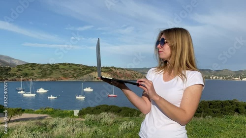 Beautiful female-traveler speaks on skype by laptop on a hill with panoramic views of the Mediterranean Sea, boats and mountains.