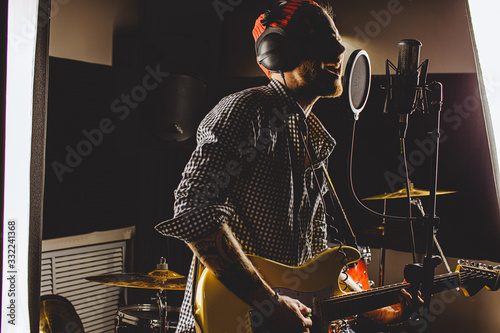young caucasian guitarist contemporaneously play guitar and sing a song in recording studio. handsome guy in red hat emotionally sing song, keen on music