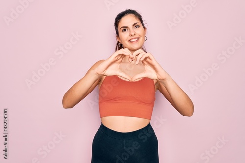 Young beautiful fitness woman wearing sport excersie clothes over pink background smiling in love showing heart symbol and shape with hands. Romantic concept.