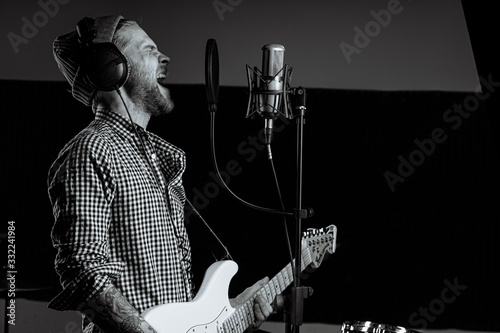 black and white photo of singing caucasian guitarist in recording studio. talented man sing on microphone and play electric guitar, wearing headsets. music concept photo
