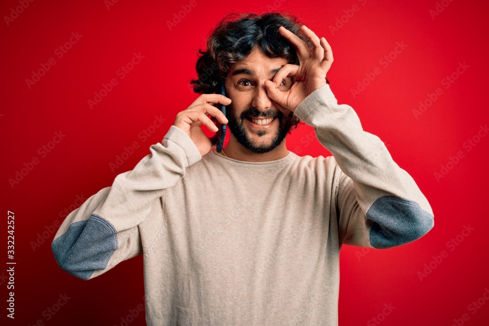 Handsome man with beard having conversation talking on the smartphone over red background with happy face smiling doing ok sign with hand on eye looking through fingers Handsome man with beard having conversation talking on the smartphone over red background with happy face smiling doing ok sign with hand on eye looking through fingers