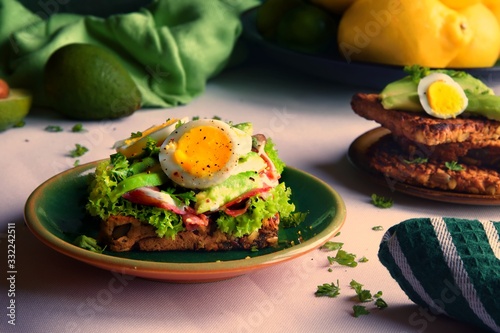 Avocado, capocollo Italian cured meat and a boiled egg slice on top of a bed of lettuce and seed loaf toast. Plate of toast sits to the right and a green dish towel in the foreground. 