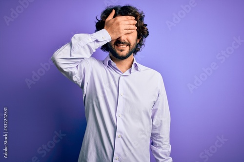 Young handsome business man with beard wearing shirt standing over purple background smiling and laughing with hand on face covering eyes for surprise. Blind concept.