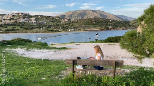 Young blond girl freelance remotely working on a laptop sitting on the bench with sea and mountain views