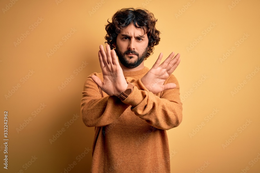 Young handsome man with beard wearing casual sweater standing over yellow background Rejection expression crossing arms and palms doing negative sign, angry face Young handsome man with beard wearing casual sweater standing over yellow background Rejection expression crossing arms and palms doing negative sign, angry face