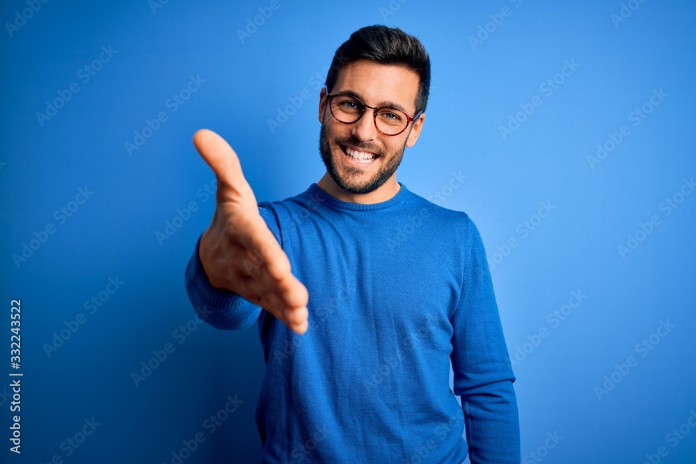 Young handsome man with beard wearing casual sweater and glasses over blue background smiling friendly offering handshake as greeting and welcoming. Successful business. Young handsome man with beard wearing casual sweater and glasses over blue background smiling friendly offering handshake as greeting and welcoming. Successful business.