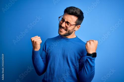 Photo Young handsome man with beard wearing casual sweater and glasses over blue background very happy and excited doing winner gesture with arms raised, smiling and screaming for success