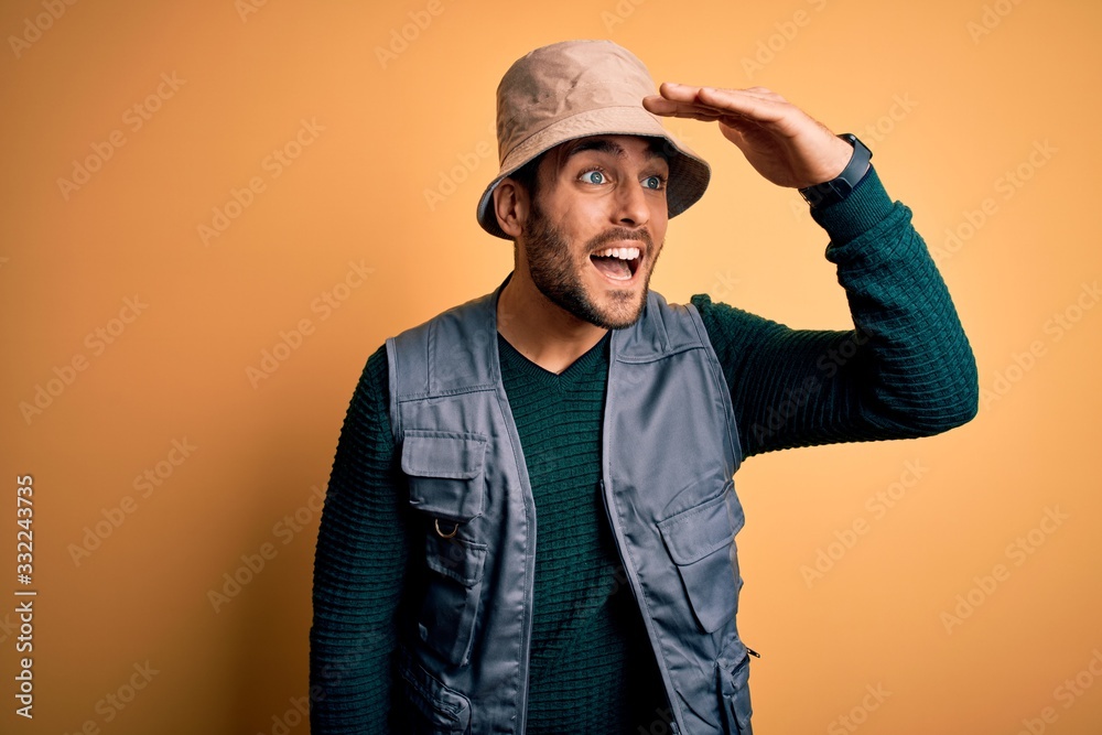 Handsome tourist man with beard on vacation wearing explorer hat over yellow background very happy and smiling looking far away with hand over head. Searching concept. Handsome tourist man with beard on vacation wearing explorer hat over yellow background very happy and smiling looking far away with hand over head. Searching concept.