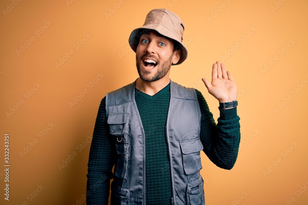 Handsome tourist man with beard on vacation wearing explorer hat over yellow background Waiving saying hello happy and smiling, friendly welcome gesture Handsome tourist man with beard on vacation wearing explorer hat over yellow background Waiving saying hello happy and smiling, friendly welcome gesture