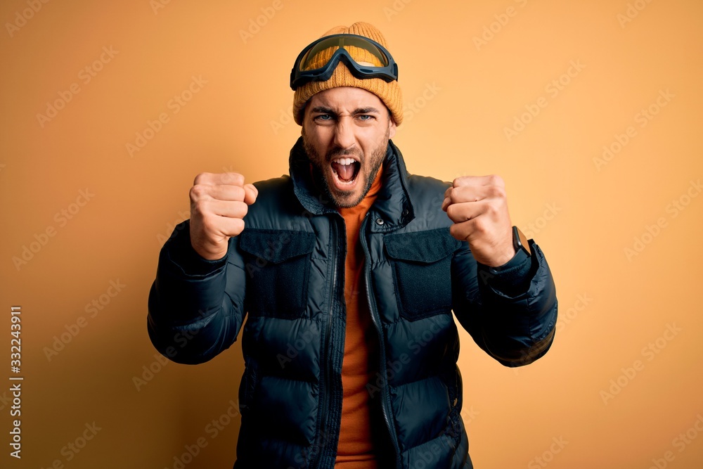 Young handsome skier man with beard wearing snow sportswear and ski goggles angry and mad raising fists frustrated and furious while shouting with anger. Rage and aggressive concept. Young handsome skier man with beard wearing snow sportswear and ski goggles angry and mad raising fists frustrated and furious while shouting with anger. Rage and aggressive concept.